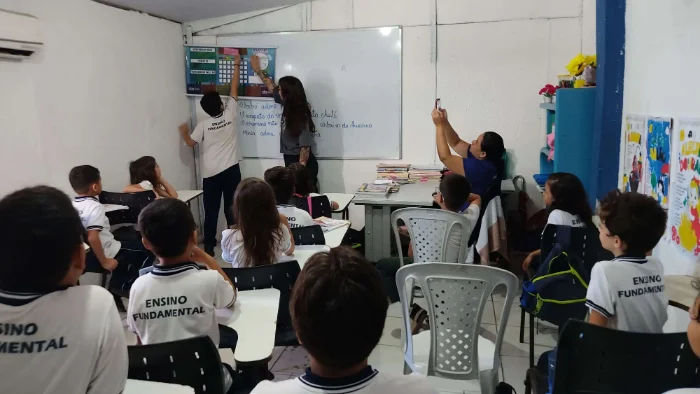 Sala de aula do Ensino Fundamental com alunos sentados e uma professora ao quadro